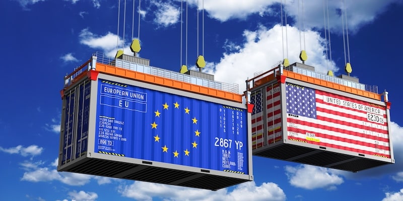 EU-US Trade Conflict Two shippings container in a blue sky filled with clouds. One container holds the EIU flag and the other the USA Flag painted on them